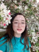 Jacquelyn is outside in front of a blossoming tree. She is wearing a teal shirt and pink glasses. She has dark, long hair.