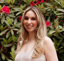 Caitlin Maloney smiles, wearing a white dress and stands in front of a blossoming rhododendron 