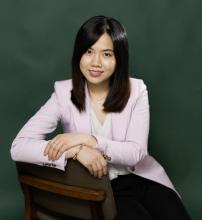 I-ling is seated in front of a dark green background on a backwards facing chair with her arms over the back. She is wearing a pink blazer and white shirt.