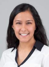 Joyti is seated in front of a white background, smiling at the camera. She is wearing a white shirt with a black collar and has long dark hair.