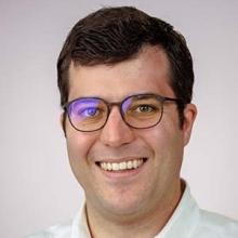 JT is centered in the photo in front of a white background. He is smiling and wearing glasses and a white button up shirt.