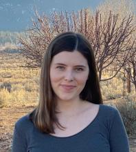 Kelsey is in front of an outdoor, scenic background. She has dark, shoulder length hair.