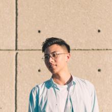 Jiachen is outdoors in front of a tan wall. They are wearing a blue collared shirt unbuttoned, a white t-shirt, and glasses.