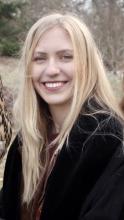 Gracia is in front of an outdoor background. She is wearing a black coat and has long, blonde hair.