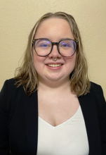 Kelsey is in front of a tan background. They are wearing a black blazer, white shirt, and glasses. They have shoulder length blonde hair.