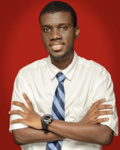 Ibrahim is in front of a red background, standing with his arms crossed. He is wearing a white button up and blue striped tie.