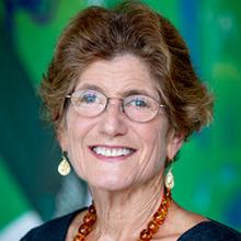 Margaret is centered in fame with a blurred, green background. She is wearing glasses, earrings, and a orange necklace with a black shirt.