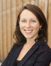 Kate is centered in front of a wood paneled background. She is medium length brown hair and is wearing a black blazer.