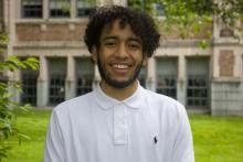 Madison is outside standing in front of a green lawn and building. He is wearing a white polo and smiling at the camera. He has short, curly hair.